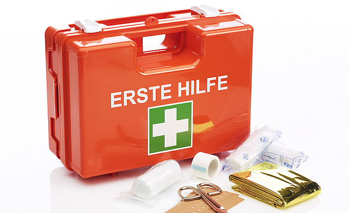Roter Koffer mit weißer Schrift "Erste Hilfe" und  darunter das weiß-grüne Symbol als Kreuz. Vor dem Koffer liegen Verbandsmaterial und eine Schere aus.