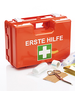 Roter Koffer mit weißer Schrift "Erste Hilfe" und  darunter das weiß-grüne Symbol als Kreuz. Vor dem Koffer liegen Verbandsmaterial und eine Schere aus.
