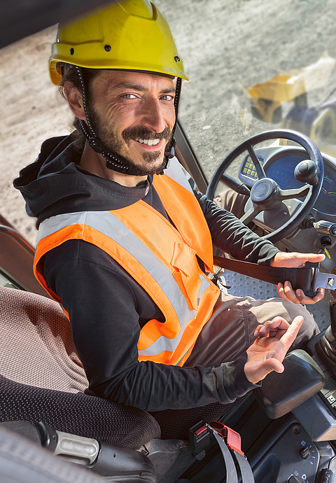 Ein Bauarbeiter deutet mit dem Finger auf den Sicherheitsheitgurt. Er sitzt in einer Baumaschine.
