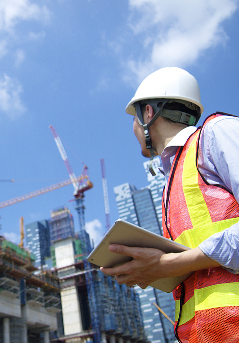 Ein Bauleiter steht mit einem Tablet vor einer Großbaustelle und sieht nach oben zu mehreren Kränen.
