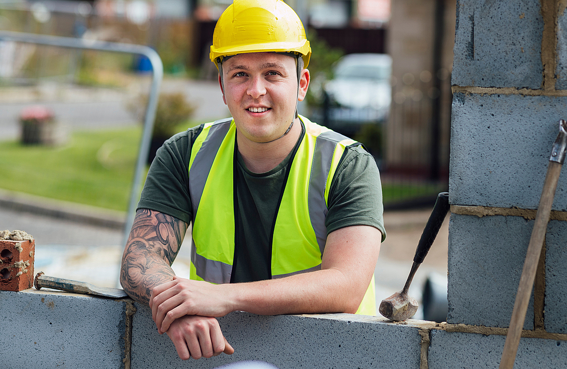 Ein junger Auszubildender im Mauerergewerk steht lächelnd in persönlicher Schutzausrüstung an eine Mauer gelehnt. 