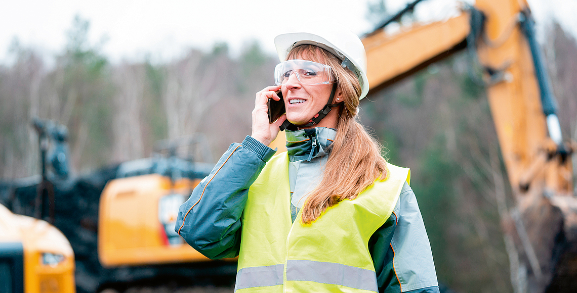 Frau in Warnweste telefoniert auf einer Baustelle mit Bagger.
