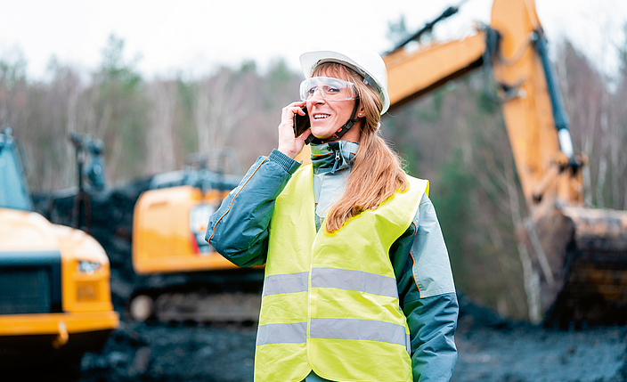 Frau in Warnweste telefoniert auf einer Baustelle mit Bagger.