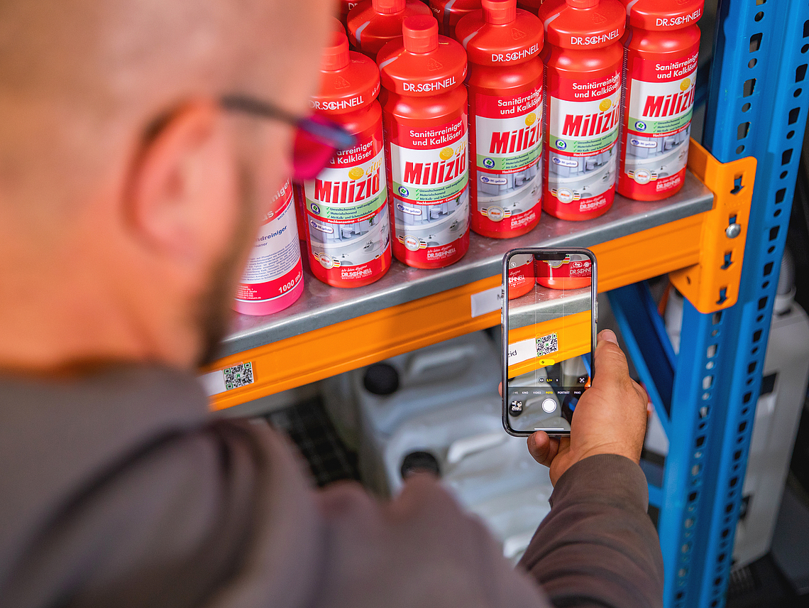 Mann scannt mit einem Smartphone einen QR-Code am Regal eines Materiallagers.
