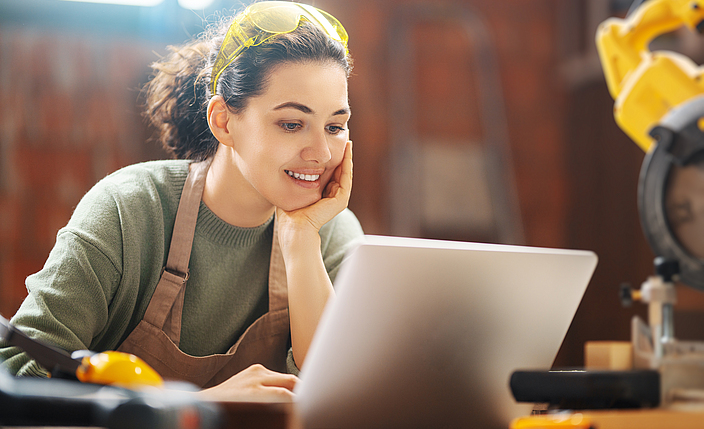 Eine junge Frau sitzt während der Arbeit mit Schutzbrille am Laptop. Sie ist in einem Werkraum. 