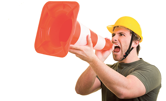 Ein Arbeiter hält einen weiß-orangenen Pylon umgekehrt in der Hand und ruft hinein.