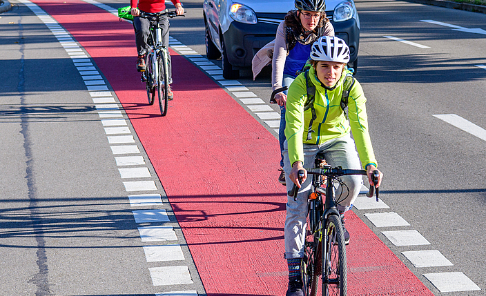 Drei Fahrradfahrer mit Helm fahren auf einem Radweg. Ein wartendes Auto möchte rechts abbiegen. 