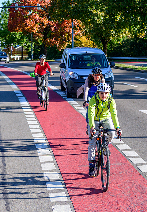 Drei Fahrradfahrer mit Helm fahren auf einem Radweg. Ein wartendes Auto möchte rechts abbiegen. 
