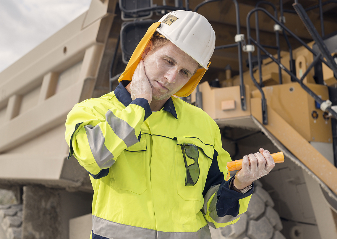 Ein Mann mit Helm und Nackenschutz cremt sich während Arbeiten im Freien auf einer Baustelle mit Sonnenschutzcreme ein.
