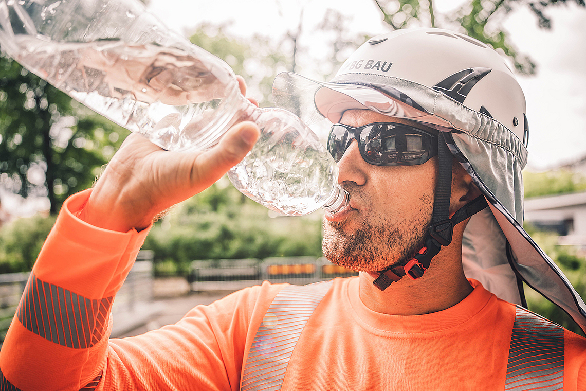 Ein Bauarbeiter mit Schutzhelm, Sonnenbrille und Nackentuch trinkt Wasser in der Pause.
