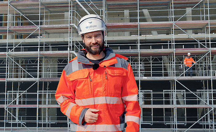 Eine Aufsichtsperson steht vor einem Gerüst. Im Hintergrund ist eine weitere Person auf einem Gerüst zu erkennen. 