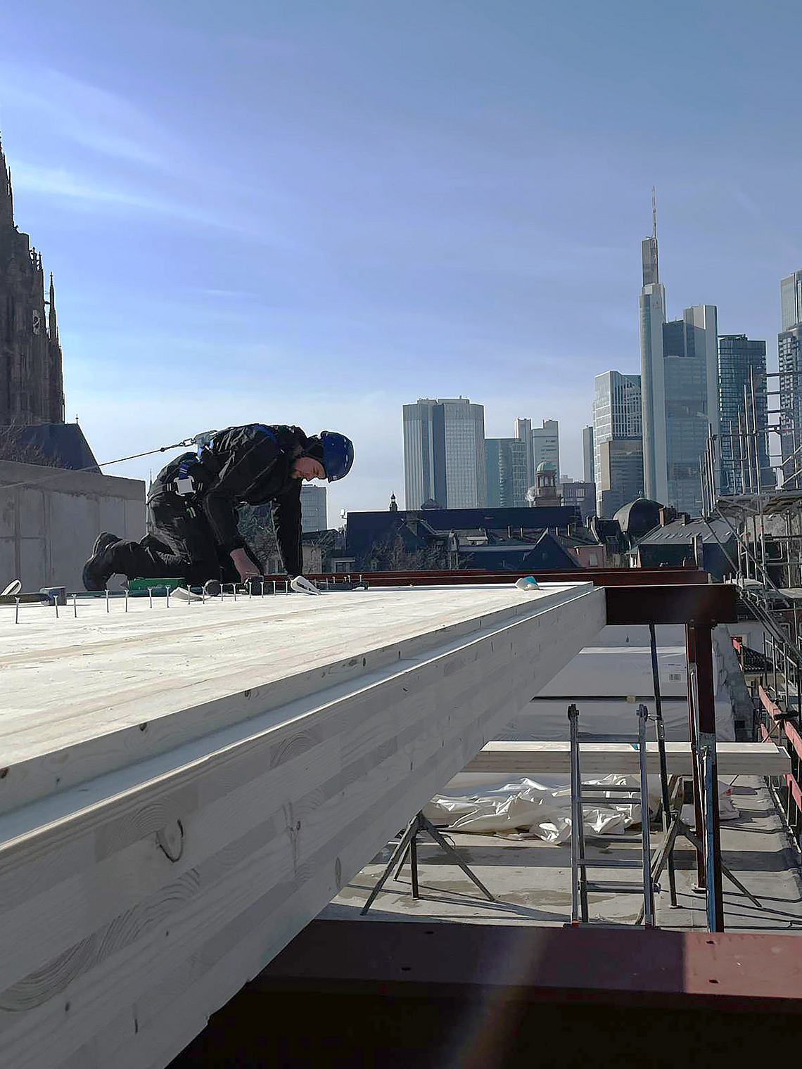Mit Lifeline gesicherter Zimmerer schlägt Nägel auf einem Bodenelement aus Holz vor dem Hintergrund der Frankfurter Skyline ein. 