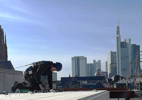 Mit Lifeline gesicherter Zimmerer schlägt Nägel auf einem Bodenelement aus Holz vor dem Hintergrund der Frankfurter Skyline ein. 