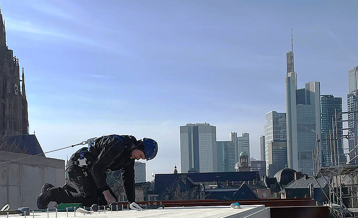 Mit Lifeline gesicherter Zimmerer schlägt Nägel auf einem Bodenelement aus Holz vor dem Hintergrund der Frankfurter Skyline ein. 