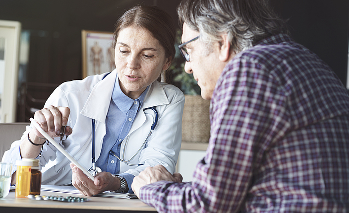 Eine Ärztin sitzt mit einem Patienten an einem Tisch. Die Ärztin zeigt dem Patienten etwas auf einem Zettel.