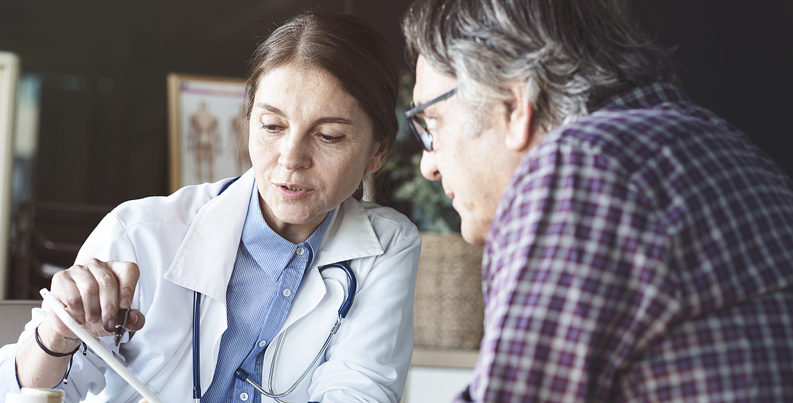 Eine Ärztin sitzt mit einem Patienten an einem Tisch. Die Ärztin zeigt dem Patienten etwas auf einem Zettel. 