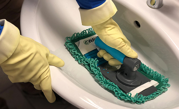Ein Waschbecken wird mit einem Reinigungsmopp gesäubert. Die Person trägt hellgelbe Schutzhandschuhe.
