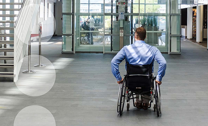 DGUV Information 215-123: Inklusion im Betrieb - Handlungshilfe für kleine und mittlere Unternehmen. Auf dem Titelbild: Ein Mitarbeiter im Rollstuhl befindet sich im Foyer eines Unternehmens,  auf dem Weg zum Fahrstuhl.
