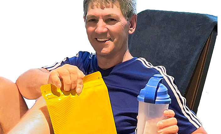 Alexander Klein auf der Trainingsbank. In einer Hand hält er einen Beutel Proteinpulver und in der anderen Hand eine Trinkflasche.