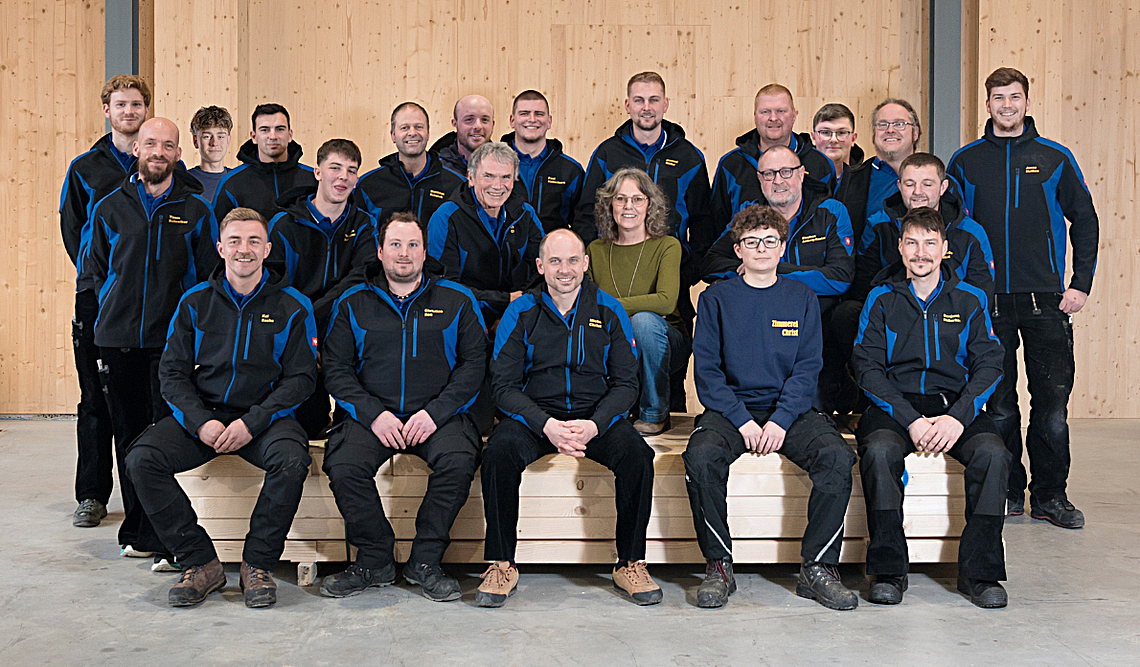 Gruppenbild aller Mitarbeiter der Zimmerei Christ vor einer Holzwand. 