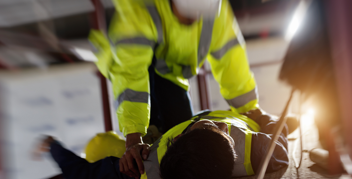 Ein Bauarbeiter leistet einem anderen Bauarbeiter bei einem Arbeitsunfall Erste Hilfe.
