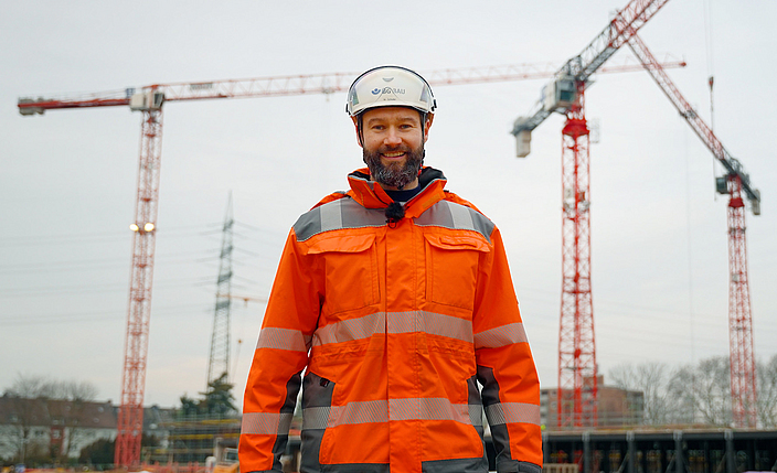 Titelbild des Videos "9 lebenswichtige Regeln im Hochbau": Marc Schuler steht auf einer Baustelle vor drei Kränen.