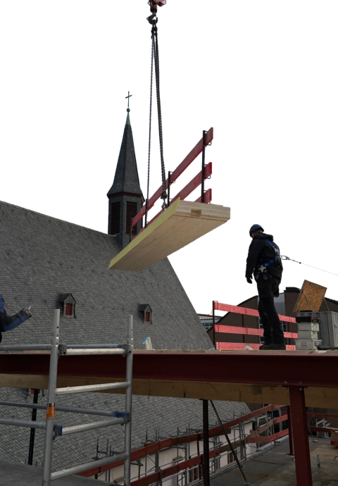 Ein mit Lifeline gesicherter Bauarbeiter erwartet ein Holzbauteil mit Seitenschutz an einem Kran. Im Hintergrund ist ein Kirchendach. 