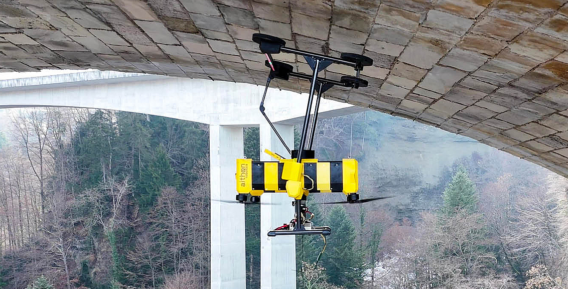Eine schwarzgelbe Drohne mit Saugfüßen hat unter einem Brückenbogen angedockt. 