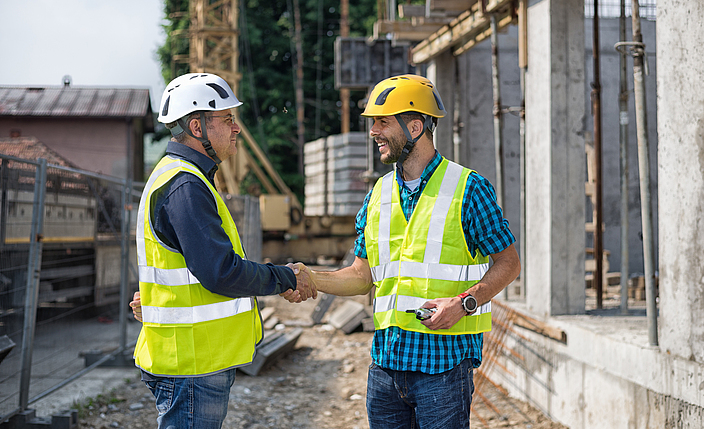 Zwei Bauleute mit Schutzhelmen und Warnwesten geben sich die Hand auf einer Baustelle.