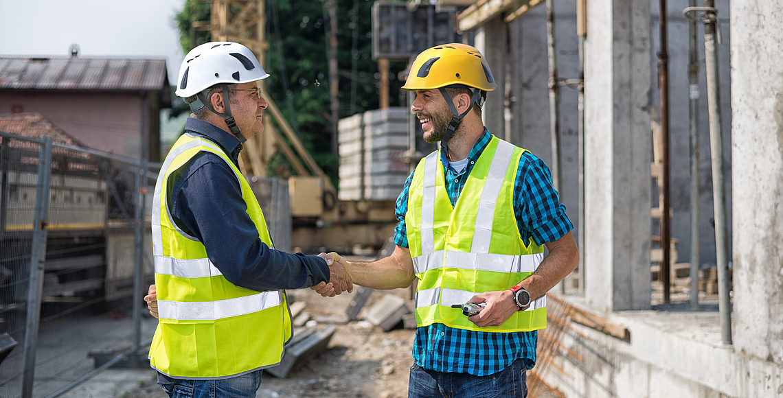 Zwei Bauleute mit Schutzhelmen und Warnwesten geben sich die Hand auf einer Baustelle. 
