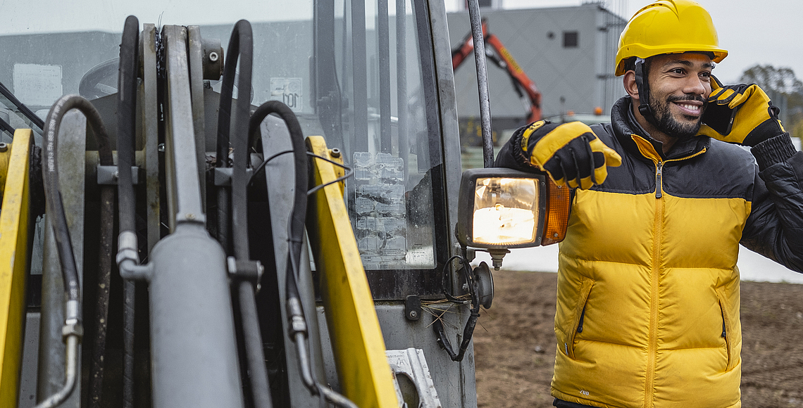 Ein Bauarbeiter telefoniert neben einem Bagger auf einer Baustelle. 