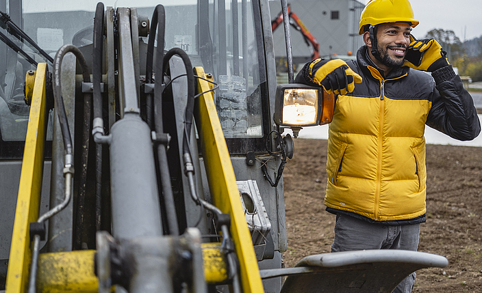 Ein Bauarbeiter telefoniert neben einem Bagger auf einer Baustelle.
