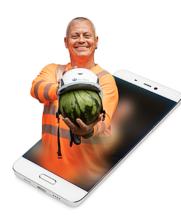 Die Aufsichtsperson Denny Hillert zeigt anhand einer Melone wie gut ein Schutzhelm den Kopf schützt. Sein Oberkörper ist auf ein Smartphone montiert.
