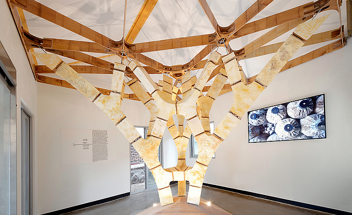 Ausstellungsraum, in dessen Zentrum ein verzweigter, künstlicher Baum aus hellen Materialien steht. Der Baum trägt die Decke des Raumes.