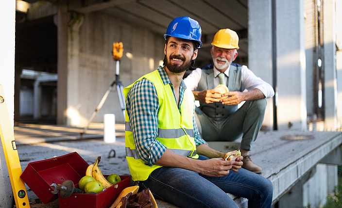 Zwei Bauarbeiter sitzen auf einer Baustelle und essen Brötchen. Sie machen Pause.