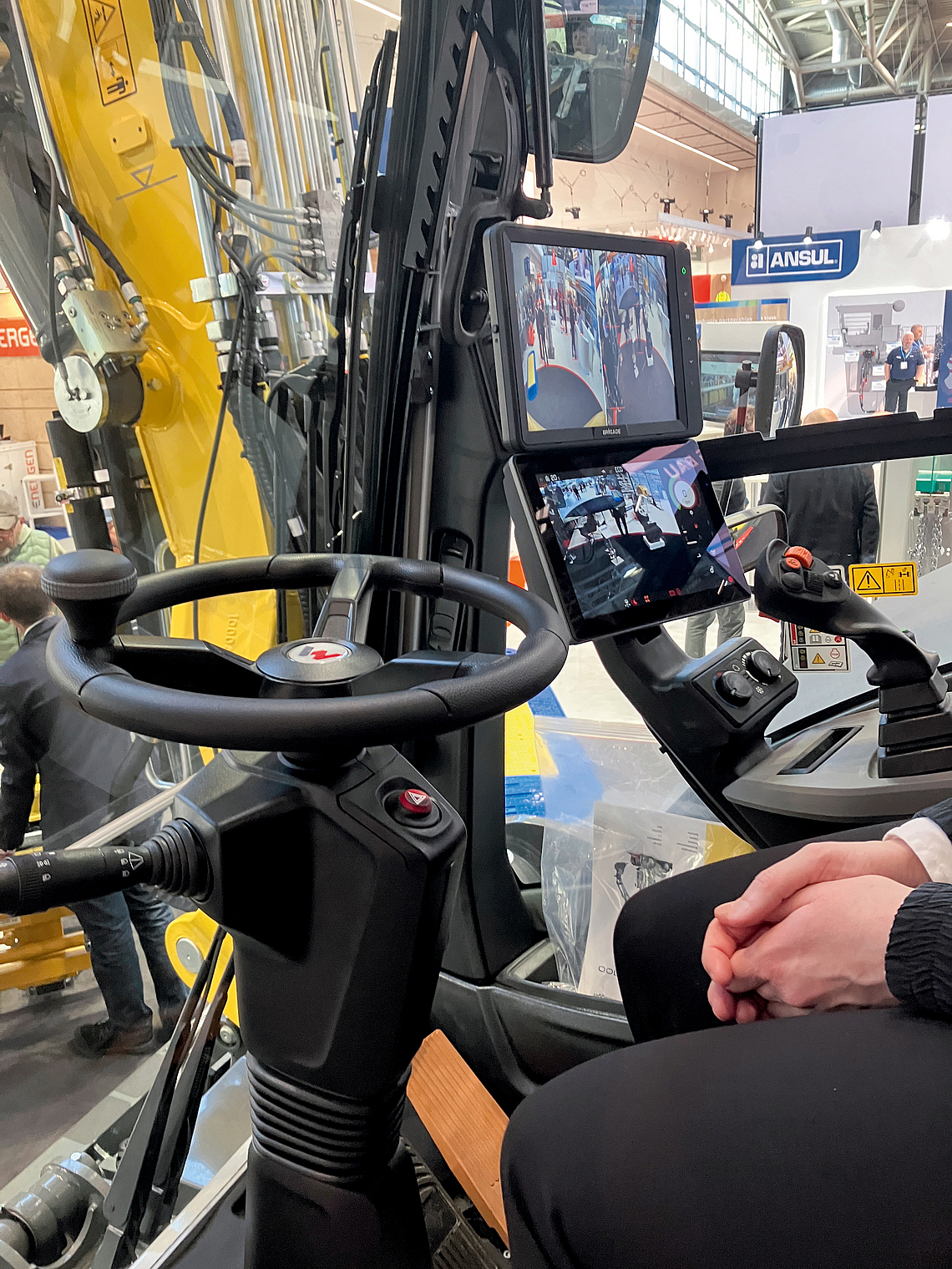 Innenaufnahme einer Baggerkabine, die in einer Messehalle aufgestellt ist. Neben dem Lenkrad sind zwei Monitore zu sehen, die das Umfeld über eine Kamera zeigen. 