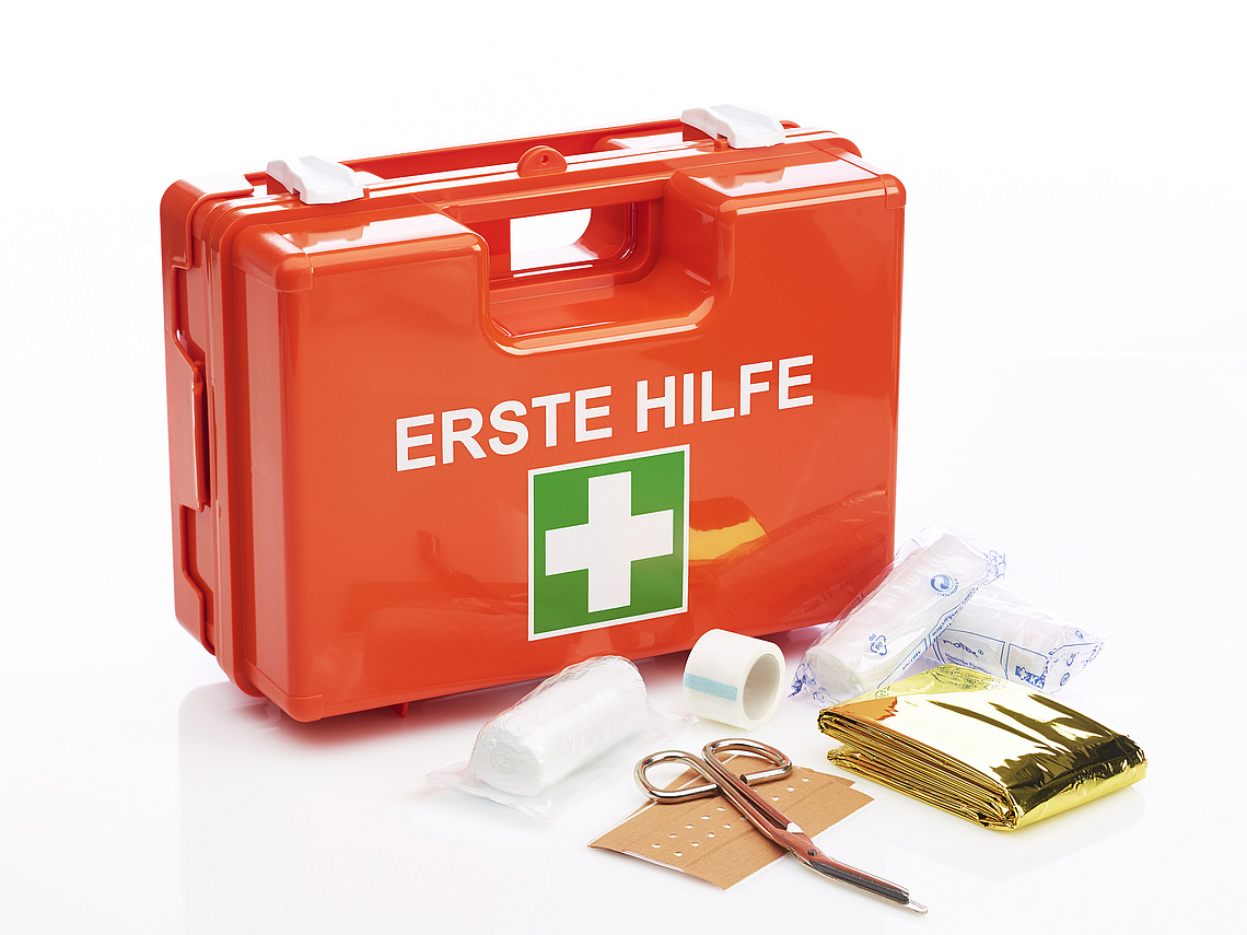 Roter Koffer mit weißer Schrift "Erste Hilfe" und  darunter das weiß-grüne Symbol als Kreuz. Vor dem Koffer liegen Verbandsmaterial und eine Schere aus.
