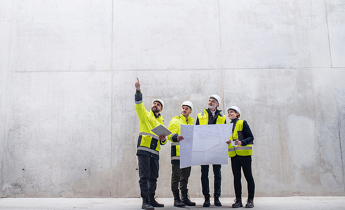 Vier Menschen in Warnkleidung und Schutzhelmen stehen mit einem Bauplan in der Hand vor einer hohen Betonwand.