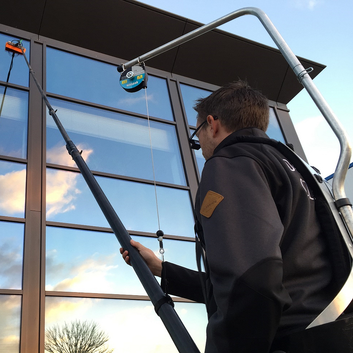 Ein Beschäftigter reinigt Fensterflächen mit einer Teleskopstange mit Bürstenaufsatz, die von einem Rucksacksystem gestützt wird.
