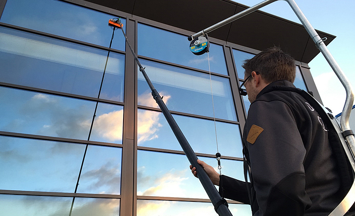 Ein Beschäftigter reinigt Fensterflächen mit einer Teleskopstange mit Bürstenaufsatz, die von einem Rucksacksystem gestützt wird.