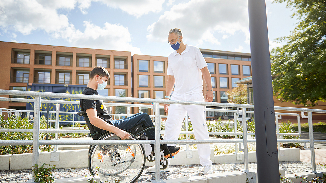 Ein Mann im Rollstuhl übt mit einem Trainer vor dem Unfallkrankenhaus Berlin.
