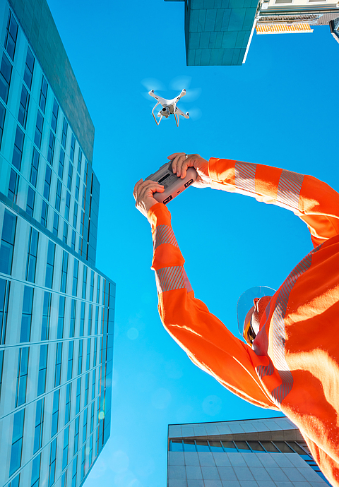 Person mit Schutzhelm und Sonnenschutz steuert eine Drohne vor einer Bürohausfassade und blauem Himmel. 