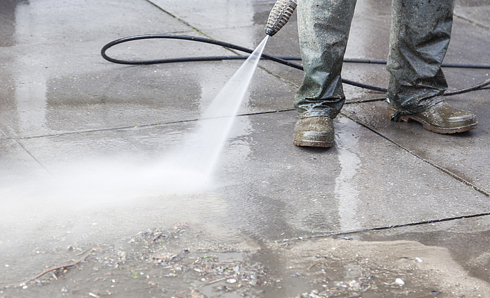 Ein Gebäudereiniger beseitigt Schmutz mit einem Hochdruck-Wasserstrahl. Dabei trägt er wasserdichte Schuhe.