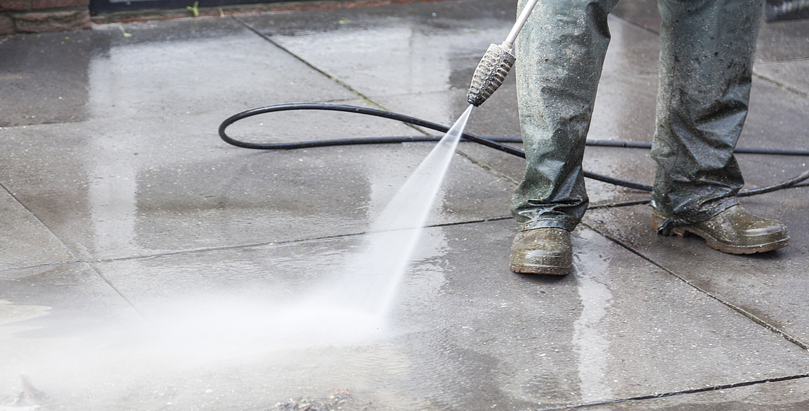 Ein Gebäudereiniger beseitigt Schmutz mit einem Hochdruck-Wasserstrahl. Dabei trägt er wasserdichte Schuhe. 