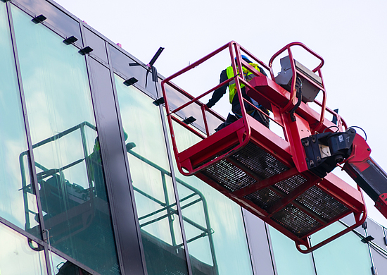 Eine Person steht auf einer Arbeitsplattform auf einem Kran und prüft eine Glasfassade.
