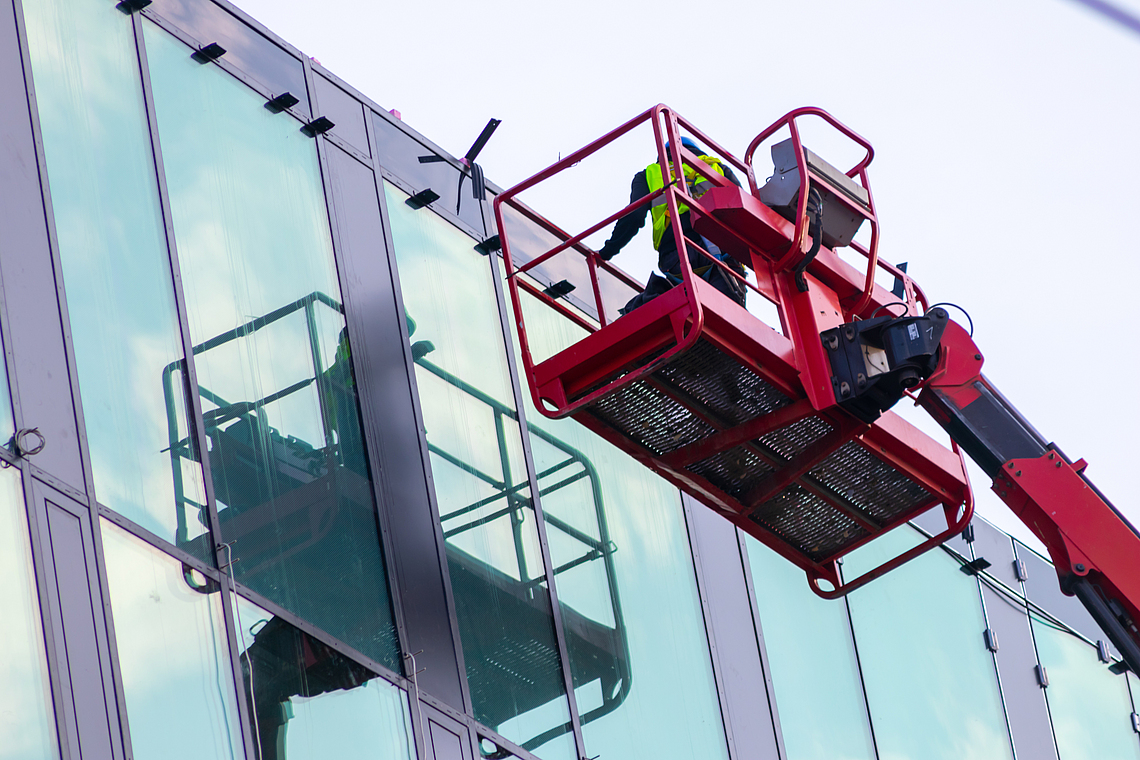 Eine Person steht auf einer Arbeitsplattform auf einem Kran und prüft eine Glasfassade.
