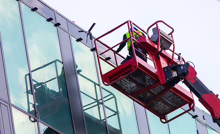 Eine Person steht auf einer Arbeitsplattform auf einem Kran und prüft eine Glasfassade.