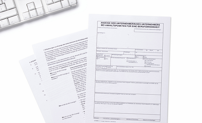 Stift und Formular für eine Verdachtsmeldung auf eine Berufskrankheit liegen neben einer Tastatur.
