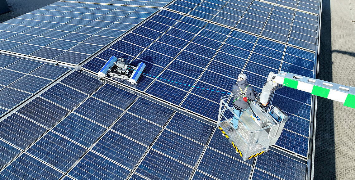 Ein Roboter reinigt eine Photovoltaikanlage auf einem Dach. Eine Person im Hubsteiger überwacht den Roboter. 