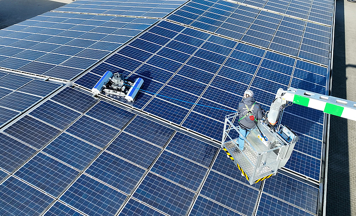 Ein Roboter reinigt eine Photovoltaikanlage auf einem Dach. Eine Person im Hubsteiger überwacht den Roboter.